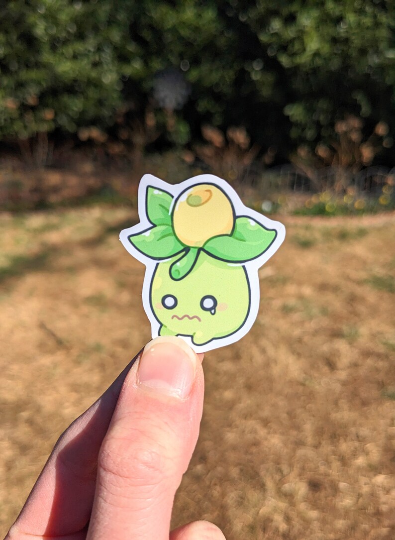 May include: A die-cut sticker featuring a cartoon character with a green body, green leaves, and a yellow fruit on top. The character has a worried expression with a small tear. The sticker is held up against a blurred outdoor background.