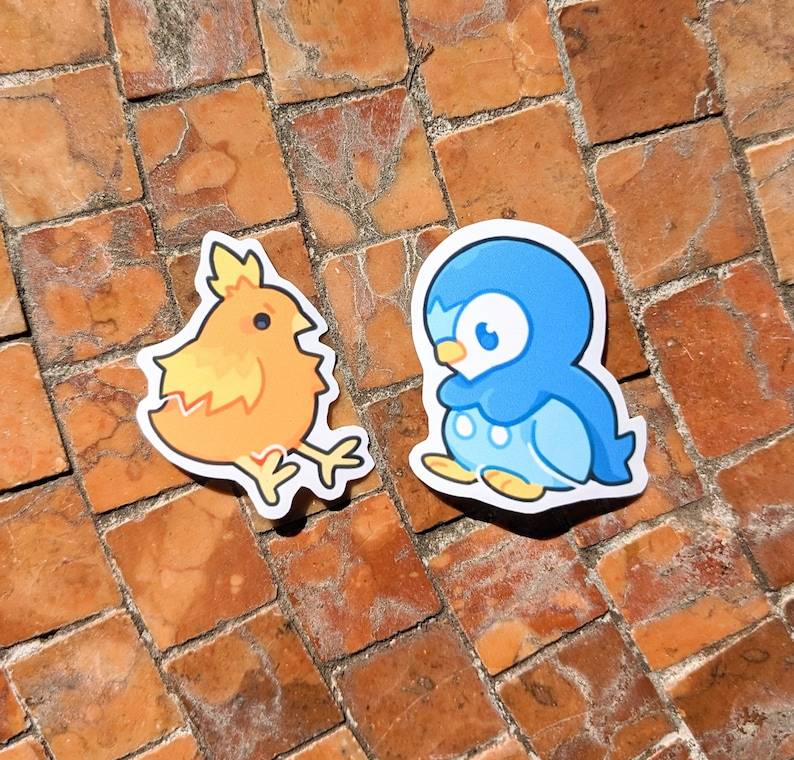 May include: Two die-cut stickers featuring cartoon characters. One is a yellow and orange chick with a jagged outline. The other is a blue penguin with white accents. Both stickers are on a brick surface.