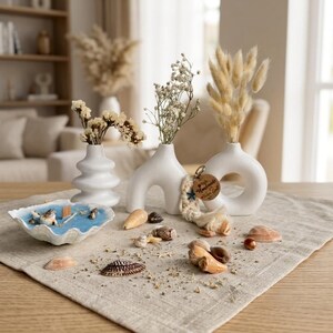 May include: Three white ceramic vases of varying shapes holding dried flowers, a shell-shaped bowl containing blue wax with a wooden wick, and scattered seashells. A small wooden tag with the text Handmade is attached to one vase.