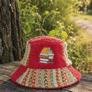 Op de afbeelding: Een rode vissershoed met een boekenontwerp. De hoed heeft een patchwork-ontwerp met een stapel boeken geborduurd op de voorkant. De rand heeft een kleurrijk, patroonontwerp. De hoed ligt op een houten oppervlak.