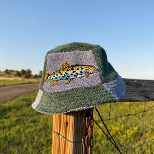 Op de afbeelding: Een bucket hat gemaakt van textuurrijke badstof met afwisselende panelen in saliegroen en lichtblauw. Het voorpaneel is voorzien van een gedetailleerde geborduurde afbeelding van een vis met gele, zwarte en blauwe schubben en vinnen.