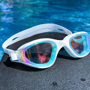 May include: White swimming goggles with a white strap and iridescent lenses rest on a dark surface. The lenses reflect a rainbow of colors. The goggles are designed for water sports and swimming.