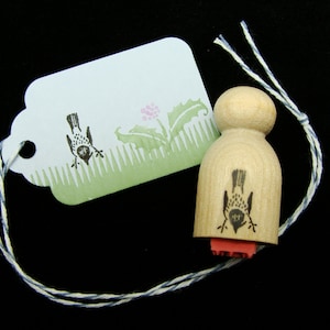 May include: A wooden stamp with a bird design. The stamp is mounted on a wooden handle and has a red ink pad. The stamp is tied to a white and blue twine.