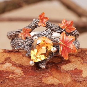 Puede incluir: Anillo de plata con una gema amarilla en forma de pera y pequeñas esferas plateadas. Adornado con ramas y hojas de arce naranja, sobre un fondo de madera. Una pieza de joyería inspirada en la naturaleza.