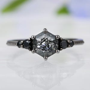 May include: A close-up of a silver ring with a hexagonal, gray gemstone at the center, featuring black inclusions. The ring has three black gemstones on each side of the central stone. The band is silver.