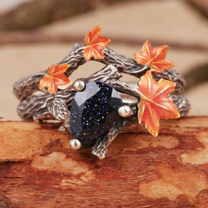Puede incluir: Anillo plateado con una gran gema azul oscuro con inclusiones brillantes. El anillo está adornado con detalles de hojas de arce naranja y un diseño similar a una rama, creando una estética inspirada en la naturaleza.