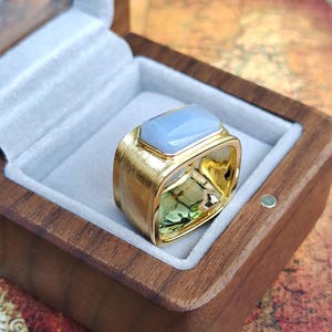 Könnte beinhalten: Ein goldfarbener Ring mit einem rechteckigen, hellblauen Edelstein, der in einem quadratischen Design gefasst ist. Der Ring wird in einer offenen Holzschatulle mit grauem Innenraum präsentiert. Die Schatulle zeigt die Details des Rings.