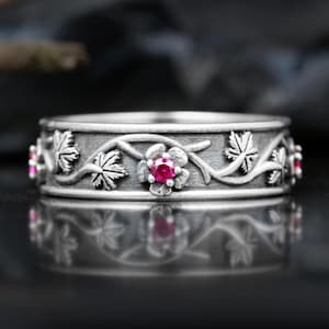 Puede incluir: Un anillo plateado con un diseño floral y de hojas. El anillo presenta una serie de hojas y flores con una pequeña gema roja y redonda en el centro de cada flor. El anillo se muestra sobre una superficie reflectante.