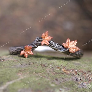 Puede incluir: Un anillo plateado con un diseño en forma de rama, adornado con tres hojas de arce color cobre. La banda del anillo se curva suavemente, y las hojas están intrincadamente detalladas, creando una estética inspirada en la naturaleza.
