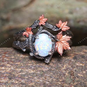 Puede incluir: Anillo de plata con una piedra de ópalo ovalada e iridiscente. El anillo presenta un diseño similar a una rama y detalles de hojas de arce de color cobre. El anillo se encuentra sobre un fondo de madera natural.