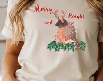 May include: A sand-colored t-shirt featuring a festive bird design. The bird is adorned with reindeer antlers and Christmas lights, perched on a bed of green foliage. The words "Merry and Bright" are written above the bird.