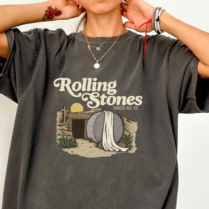 May include: Pepper-colored t-shirt with the text "Rolling Stones Since AD 33" and a graphic of a tomb with a large stone and a rising sun. The shirt has short sleeves and a relaxed fit.
