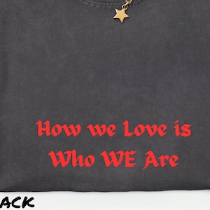 May include: A black Comfort Colors t-shirt with the text "How we Love is Who WE Are" in red. A gold star pendant necklace and a brown hat are also visible. The shirt is size XL-TG-EG.