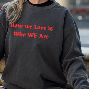 May include: Dark grey sweatshirt featuring the red text "How we love is Who WE Are". The sweatshirt has a crew neck and long sleeves. The garment appears to be made of a soft, comfortable material.