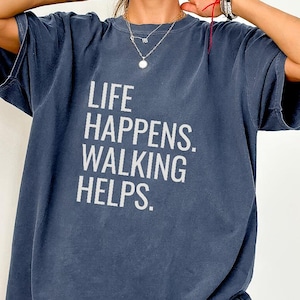 May include: A dark blue short-sleeve t-shirt featuring the white text LIFE HAPPENS. WALKING HELPS. printed on the front. The garment has a crew neckline and a relaxed fit, constructed from a solid-colored fabric with a slightly faded appearance.