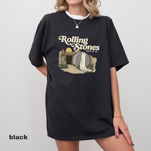 May include: Black t-shirt with the text "Rolling Stones Since AD 33" above an illustration of a tomb with a stone and a rising sun. The shirt has a white collar and a relaxed fit.