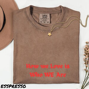 May include: A brown t-shirt with the text "How we Love is Who WE Are" in red. The shirt has a crew neck and a tag that says "COMFORT COLORS XL-TG-EG". A gold star pendant necklace and a brown hat are also in the image.