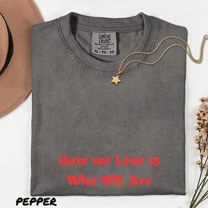 May include: A grey Comfort Colors t-shirt with the text "How we Love is Who WE Are" in red. A gold star pendant necklace rests on the shirt. The shirt is folded and displayed with a hat and flowers.