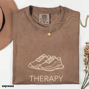 May include: A brown t-shirt featuring a line art graphic of two sneakers above the word THERAPY in a light beige color. The shirt has a Comfort Colors brand label at the inner neckline and is paired with a gold chain necklace with a star charm.