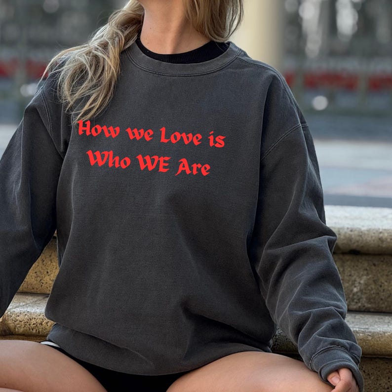May include: Dark grey sweatshirt featuring the red text "How we love is Who WE Are". The sweatshirt has a crew neck and long sleeves. The garment appears to be made of a soft, comfortable material.