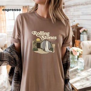 May include: Espresso-colored t-shirt featuring the text "Rolling Stones" above an illustration of an open tomb with a rising sun. The shirt has rolled-up sleeves and is paired with a plaid shirt.
