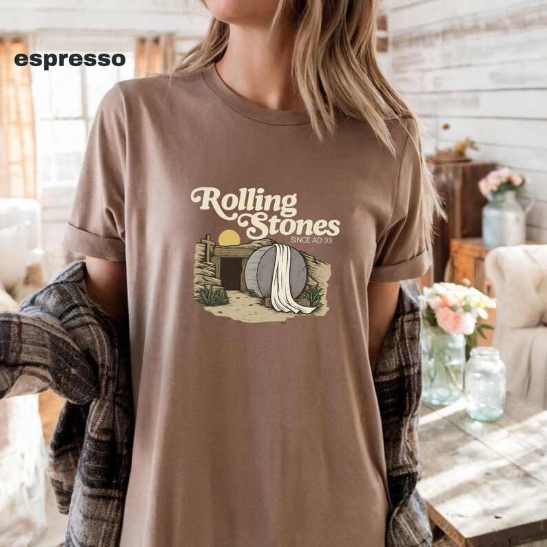 May include: Espresso-colored t-shirt featuring the text "Rolling Stones" above an illustration of an open tomb with a rising sun. The shirt has rolled-up sleeves and is paired with a plaid shirt.