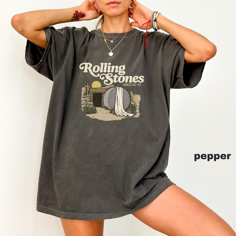 May include: Pepper-colored t-shirt with the text "Rolling Stones Since AD 33" and a graphic of a tomb with a large stone and a rising sun. The shirt has short sleeves and a relaxed fit.