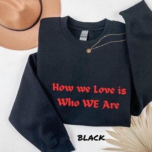 May include: Black crew neck sweatshirt with the red text "How we Love is Who WE Are". The word "BLACK" is printed at the bottom. A gold necklace and a brown hat are also visible.