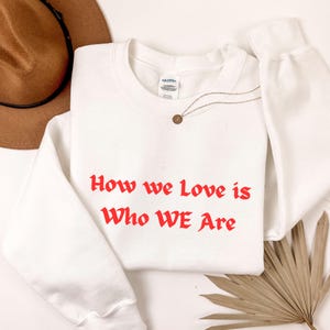 May include: A white sweatshirt with the red text "How we Love is Who WE Are." A gold-coloured necklace with a small pendant is draped across the sweatshirt. A brown hat is visible in the background.