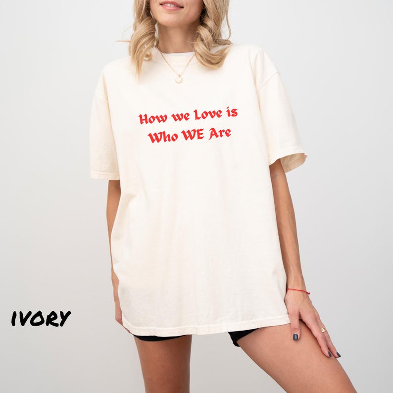 May include: Ivory-colored t-shirt with the text "How we Love is Who WE Are" in red. The shirt has short sleeves and a relaxed fit. The word "IVORY" is printed in black at the bottom left.