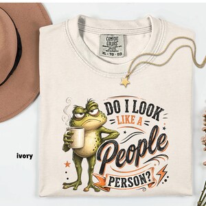 Pode incluir: Uma t-shirt cor de marfim com um gráfico de um sapo verde rabugento segurando uma caneca de café fumegante. O texto diz "DO I LOOK LIKE A PEOPLE PERSON?". A t-shirt tem uma etiqueta Comfort Colors no pescoço, tamanho XL, feita de algodão.
