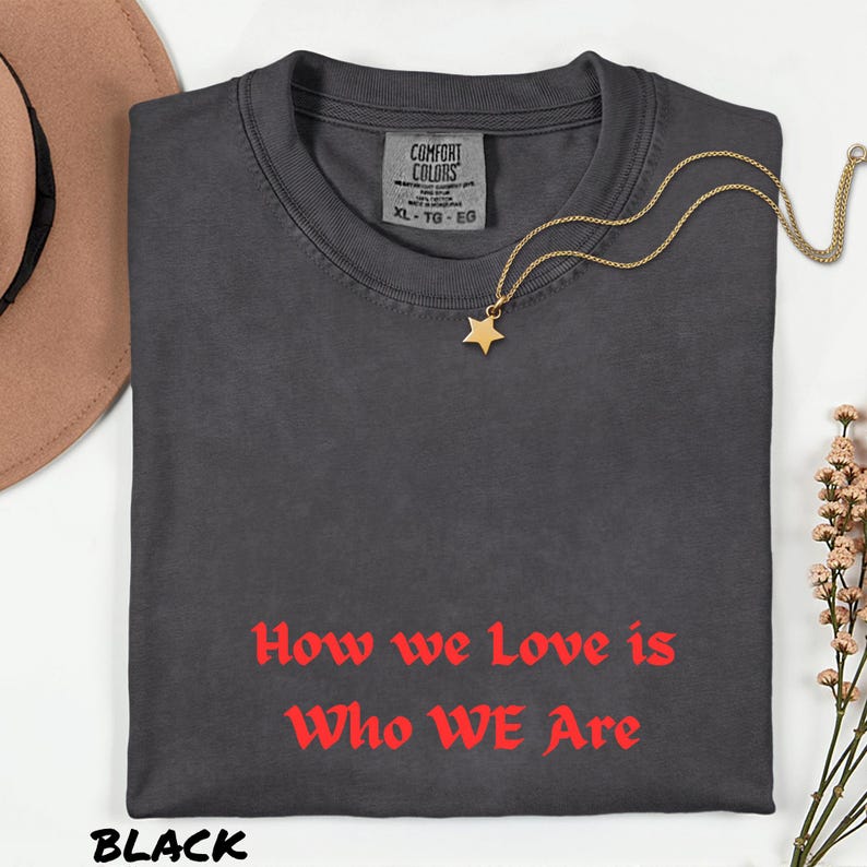 May include: A black Comfort Colors t-shirt with the text "How we Love is Who WE Are" in red. A gold star pendant necklace and a brown hat are also visible. The shirt is size XL-TG-EG.