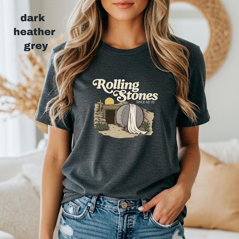 May include: Dark heather grey t-shirt featuring the text "Rolling Stones Since AD 33" above an illustration of a tomb with a large stone and a rising sun. The shirt is a short-sleeved crew neck.