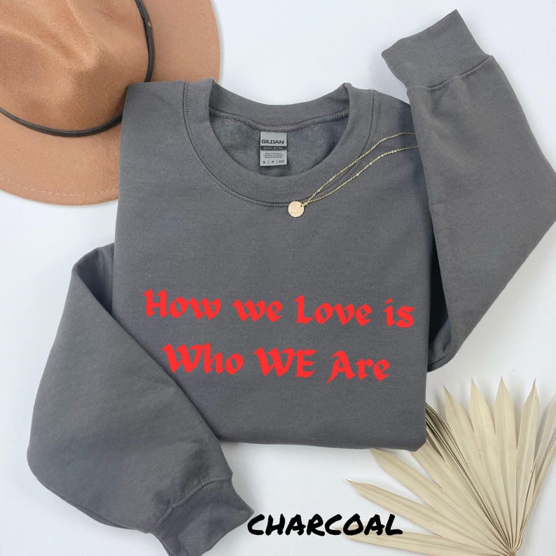 May include: A charcoal grey sweatshirt with the red text "How we Love is Who WE Are". A gold necklace with a pendant rests on the garment. The word "CHARCOAL" is printed at the bottom of the image.