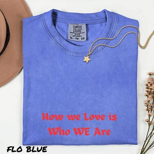 May include: A blue XL Comfort Colors t-shirt with the text "How we Love is Who WE Are" in red. A gold star pendant necklace is draped on the shirt. The shirt is labelled "FLO BLUE".