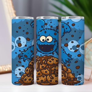 May include: A set of three blue stainless steel tumblers featuring a cartoon character with a blue furry body, large eyes, and a wide grin. The character is surrounded by chocolate chip cookies and cookie crumbs. The tumblers are designed for cold beverages.