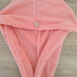 Pode incluir: Uma toalha de cabelo de microfibra cor-de-rosa com capuz. A toalha é presa com um pequeno botão de madeira em forma de rosto de urso. A toalha foi concebida para envolver a cabeça e absorver a água após a lavagem do cabelo.