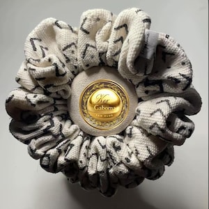 Puede incluir: Una goma para el pelo blanca con un patrón de zigzag negro. La goma para el pelo tiene un emblema dorado en el centro con el texto "Ker Gaboma FAN CLUB". Una pequeña etiqueta con "Ker Gaboma" está adjunta.