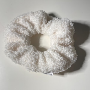 Puede incluir: Una esponjosa y blanca goma para el pelo con una apariencia texturizada similar a la felpa. El accesorio para el cabello es circular con un agujero en el centro. Una pequeña etiqueta con texto es visible en el lateral.