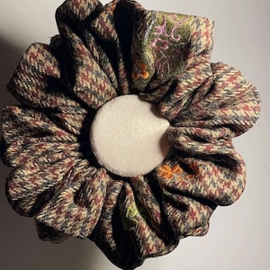 Puede incluir: Una gran goma para el pelo con forma de flor. La goma está hecha de una tela de tweed estampada en tonos marrones, rojos y verdes. El centro es de tela redonda blanquecina. Un pétalo tiene detalles florales bordados en rosa y naranja.