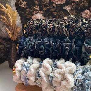 Puede incluir: Una pila de coleteros en varios patrones y colores. El coletero superior tiene un ribete dorado y un diseño de estampado de leopardo. El coletero del medio es azul marino y verde azulado. El coletero inferior es blanco con un estampado floral azul.