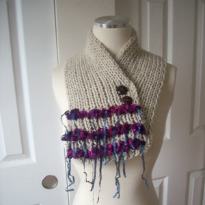 May include: A cream-colored knitted scarf with a purple and blue ribbon trim. The scarf is folded in half and has a button closure.