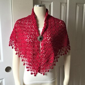 May include: A red crocheted shawl with a decorative beaded fringe. The shawl is displayed on a white mannequin.