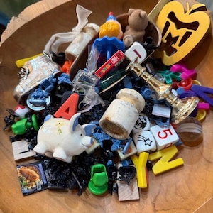 May include: A wooden bowl filled with a variety of small objects. Items include a white pig figurine, dice, letters, a small teddy bear, a gold-colored apple-shaped object, and other miniature toys and trinkets.