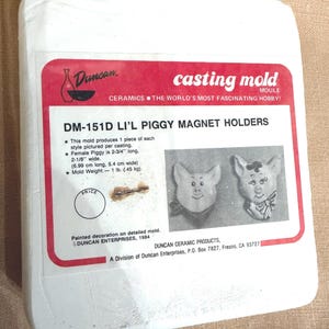 May include: A white casting mold box with a red label featuring the text "casting mold" and "DM-151D LI'L PIGGY MAGNET HOLDERS." The label includes images of two pig-shaped magnet holders. The mold produces 1 piece of each style. The female piggy is 2-3/4" long.