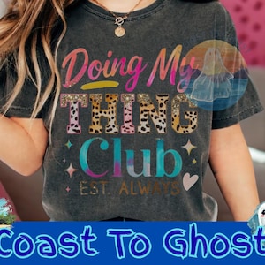 Könnte beinhalten: Dunkelgraues T-Shirt mit dem Text "Doing My Thing Club" in einer bunten, glitzernden Schrift. Das Wort "THING" hat ein Leopardenmuster. Das Shirt zeigt auch Sterne, Herzen und die Worte "EST. ALWAYS."