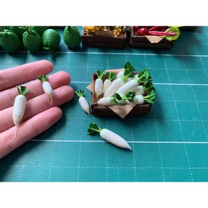 May include: Miniature white radishes with green tops are displayed in a small wooden crate and held in a hand. Other miniature produce, including green limes, are visible in the background. The scene is set on a green surface.