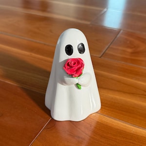 Puede incluir: Figura de fantasma blanco con ojos ovalados negros, sosteniendo una rosa roja con hojas verdes. El fantasma est&aacute; envuelto en una s&aacute;bana blanca. El objeto es decorativo.