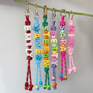 May include: Colorful beaded keychains spelling out names: EMILY, JAMIE, STEFAN, BETSY, SARAH, and NINA. Each keychain features a different color scheme, with gold-tone clasps and decorative knots. The beads are in various colors and shapes.