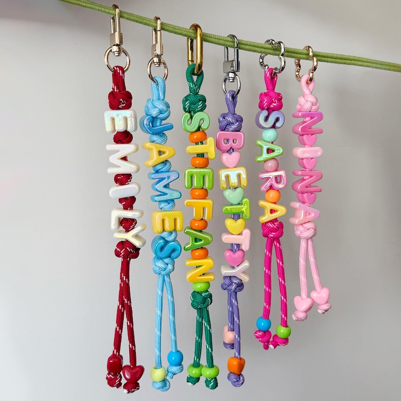 May include: Six personalised keychains hanging from a green cord, each featuring colourful plastic letter beads spelling names like Emily, James, Stefan, Betty, Sara, and Nina, attached to metal lobster clasps with knotted nylon cord accents.